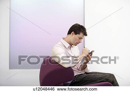 Stock Photograph - Casually dressed Businessman sitting on modern sofa, Using PDA, side view. Fotosearch