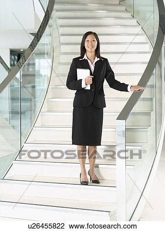 Stock Image - Confident Businesswomen holding folder, standing on stairs, hand on banister. Fotosearch
