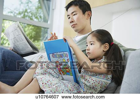 Stock Photo - Daughter with picture book, sitting beside father on sofa with newspaper, side view. Fotosearch