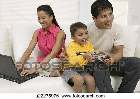 Stock Photograph - Father playing video game with son by mother using laptop on couch. Fotosearch
