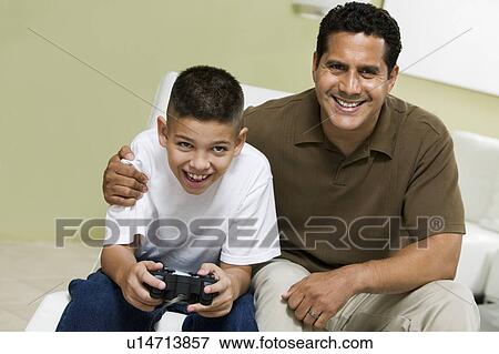Stock Photo - Father with Son Playing Video Game on sofa in living room, portrait. Fotosearch