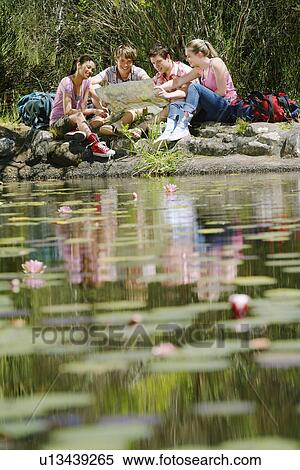 Stock Photography - Four teenagers (16-217 years) sitting by lake, reading map, selective focus. Fotosearch