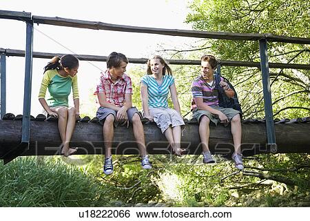 Stock Photograph - Four teenagers 916-17 years) sitting on wooden bridge, talking, smiling. Fotosearch