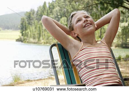 Stock Image - Girl (7-9) by lake, leaning back on deck chair.. Fotosearch