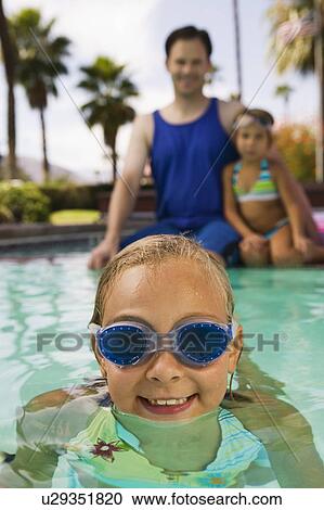 Stock Image - Girl (7-9) in swimming pool, father and sister (7-9) sitting on pool edge in background, front view portrait.. Fotosearch