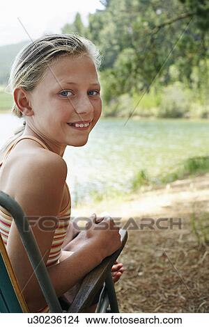 Picture - Girl (7-9) sitting on deck chair by lake.. Fotosearch
