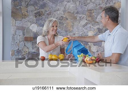 Stock Image - Husband handing wife orange from countertop. Fotosearch
