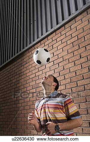 Stock Image - Man heading football by brick wall. Fotosearch