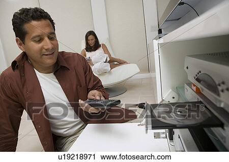 Stock Image - Man inserting DVD and using remote control in living room, close up. Fotosearch