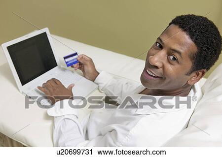 Stock Image - Man on sofa Using Credit Card to Make Purchase with Laptop, portrait, high angle view. Fotosearch