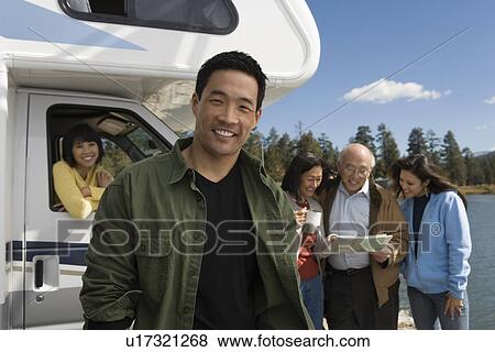 Stock Photo - Man standing in front of RV at lake with family behind. Fotosearch