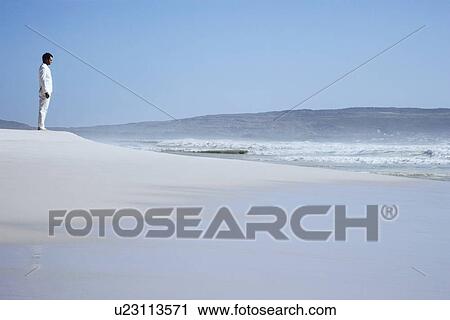 Stock Image - Man standing on sand dune on beach, side view. Fotosearch