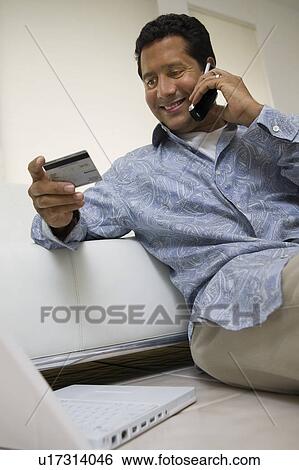 Man using mobile phone holding credit card in living room View Large Photo Image Stock Photograph - Man using mobile phone holding credit card in living room. Fotosearch