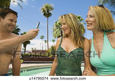Man using mobile phone, photographing two women by swimming pool. View Large Photo Image Picture - Man using mobile phone, photographing two women by swimming pool.. Fotosearch