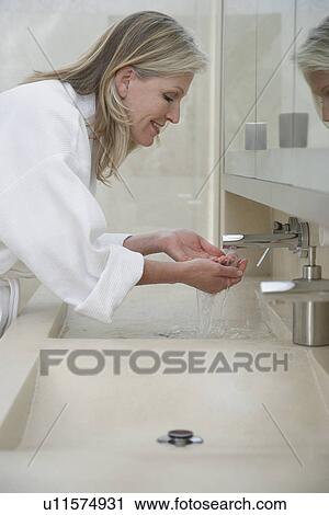Stock Image - Mature woman in bathrobe washing face over sink, profile. Fotosearch