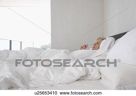 Stock Image - Mature woman lying in bed with thermometer in mouth, side view. Fotosearch