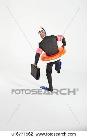 Stock Image - Middle-aged businessman in flippers, inflatable rubber ring, snorkel and goggles waddling, carrying briefcase, back view. Fotosearch