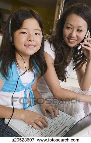 Stock Photo - Mother on cell phone watching daughter using laptop in living room, portrait. Fotosearch