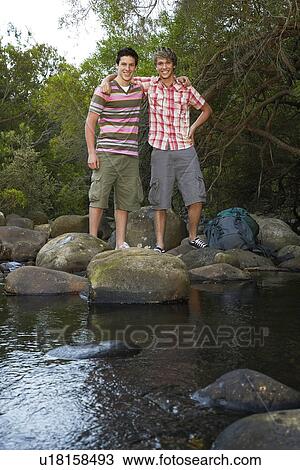 Stock Image - Portrait of two teenage boys (16-17 years) standing on stones by river, arm around, smiling. Fotosearch