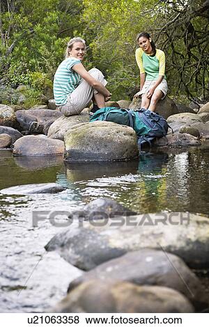 Stock Photo - Portrait of two teenage girls (16-17 years) sitting on stones by river. Fotosearch