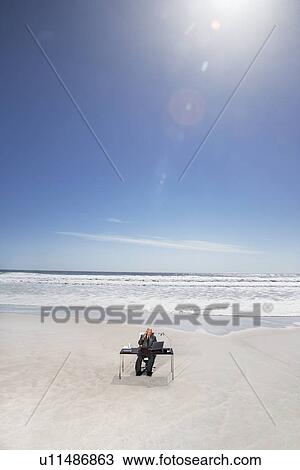 Stock Image - Senior business man sitting at office desk on beach, elevated view. Fotosearch