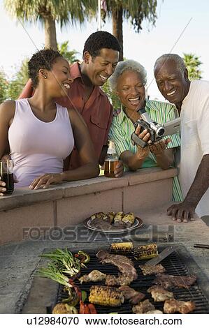 Stock Image - Senior couple and mid-adult couple looking at camcorder at outdoor barbecue.. Fotosearch