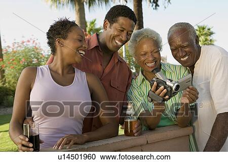 Stock Image - Senior couple and mid-adult couple looking at camcorder outdoors.. Fotosearch