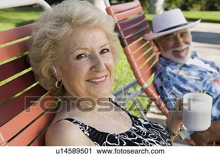 Stock Image - Senior Couple sitting on lawn chairs, woman listening to earphones and holding cup, portrait.. Fotosearch