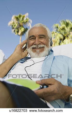 Stock Image - Senior Man sitting outdoors, listening to headphones, low angle portrait.. Fotosearch