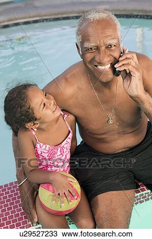 Stock Image - Senior man using mobile phone, sitting by pool with granddaughter (5-6), portrait.. Fotosearch