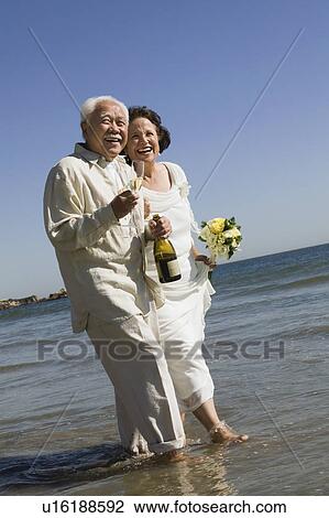 Stock Image - Senior newly weds walking in ocean. Fotosearch