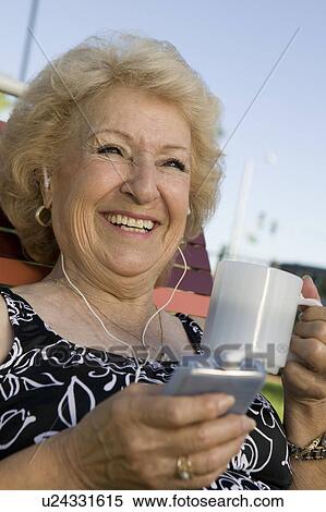 Stock Photography - Senior Woman outdoors, listening to portable music player, holding cup, smiling.. Fotosearch