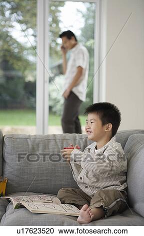 Stock Image - Smiling Boy on sitting barefooted on Sofa with Coloring Book, father in background talking on phone. Fotosearch