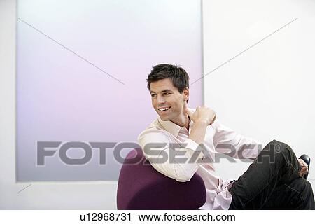 Stock Image - Smiling Businessman sitting on modern sofa, legs crossed, looking over shoulder. Fotosearch