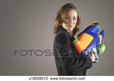 Stock Image - Smiling businesswoman standing, holding water gun, side view. Fotosearch