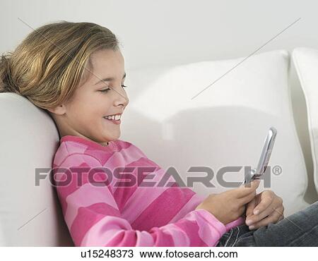 Stock Image - Smiling Girl lying on sofa, using Cell Phone, side view. Fotosearch
