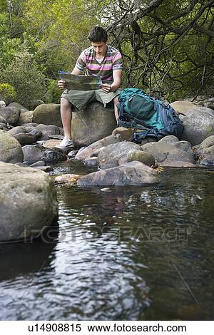 Stock Photography - Teenage boy (16-17 years) sitting on stone by river reading map. Fotosearch