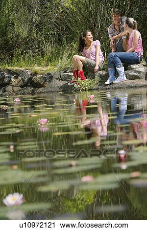 Stock Image - Teenage girls and boy (16-17 years) sitting on stones by lake, smiling. Fotosearch