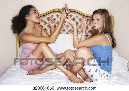 Stock Photograph - Teenage Girls sitting legs crossed on bed, looking in compacts, applying make-up, side view. Fotosearch