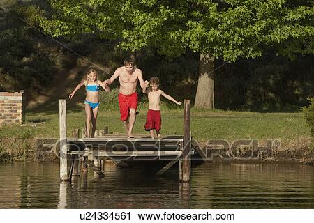 Stock Image - Two children (7-9) holding hands with father, jumping into lake.. Fotosearch