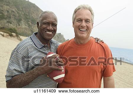 Stock Photo - Two men with American football on beach, (portrait). Fotosearch