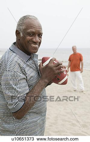 Stock Image - Two senior men playing american football on beach. Fotosearch