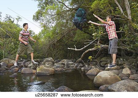 Stock Photo - Two teenage boys (16-17 years) standing on stones by river, throwing backpack. Fotosearch