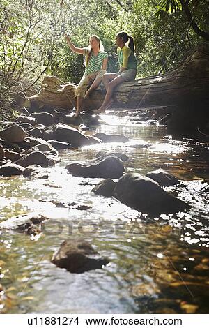 Picture - Two teenage girls (16-17 years) sitting on tree trunk by stream. Fotosearch