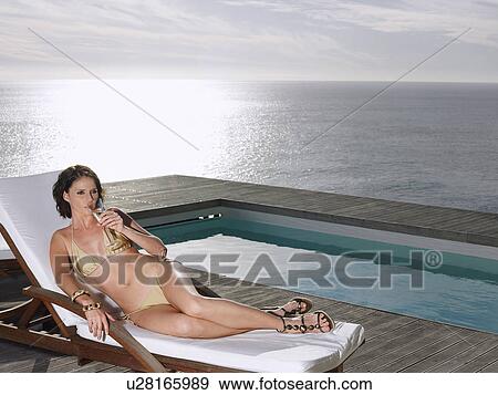 Stock Photo - Woman lying on deck chair sipping champagne at poolside by ocean. Fotosearch