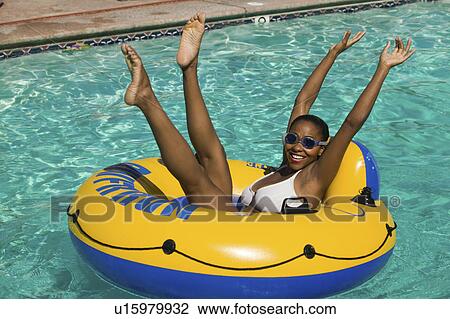 Stock Image - Woman lying on inflatable raft in swimming pool with arms and legs raised, portrait.. Fotosearch