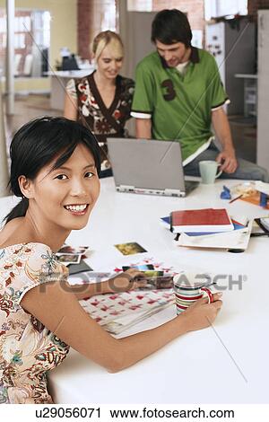 Stock Image - Woman sitting at table with two people using laptop in background.. Fotosearch