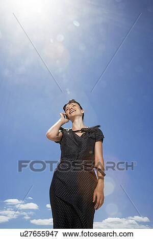 Stock Photo - Woman using mobile phone standing against sky, low angle view. Fotosearch
