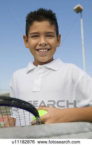 Stock Photograph - Young boy with tennis racket and ball at net on tennis court, portrait, close up. Fotosearch