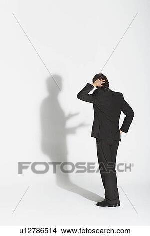 Young businessman standing, hand on head and hip, arguing with own shadow View Large Photo Image Picture - Young businessman standing, hand on head and hip, arguing with own shadow. Fotosearch